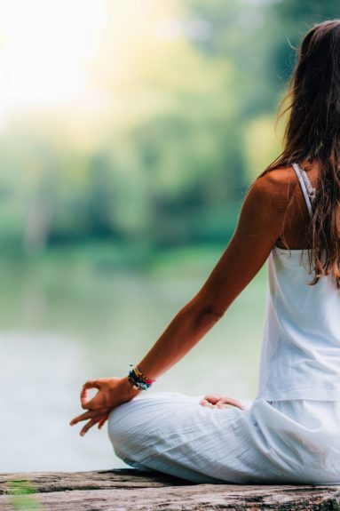 Woman doing yoga by the lake, sitting in lotus pose with fingers in mudra position.